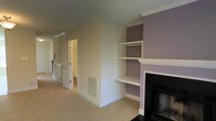 This two-story home offers a thoughtfully designed layout with classic architectural details and functional living spaces. The main living area is a standout feature, centered around a traditional fireplace with a crisp white mantel and sleek black surround, creating a warm and inviting focal point. Adjacent to the fireplace, built-in open shelving set against a soft lavender accent wall provides both display space and a touch of character, adding visual interest to the room. Crown molding runs along the ceiling, lending a refined, classic finish to the space. Soft, neutral-toned carpeting extends throughout, contributing to a comfortable and cohesive atmosphere. The room connects seamlessly to a hallway that leads further into the home, with a staircase providing access to the second floor. A glimpse of an adjoining room hints at additional functional spaces beyond. Ample wall venting ensures consistent climate control throughout. The overall ambiance is calm and understated, with a blend of neutral creams and muted lavender tones that give the home a serene, livable quality well-suited for everyday comfort.