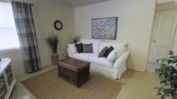 This single-floor space offers a calm and inviting atmosphere defined by a neutral color palette and thoughtfully chosen furnishings. The living area serves as the heart of the space, centered around a classic white slipcovered sofa accented with dark charcoal throw pillows, creating a crisp contrast that anchors the room. A rich, dark woven rattan coffee chest sits atop a natural jute area rug, adding warmth and texture to the light-toned tile flooring beneath. The walls are finished in a soft, warm white that enhances the airy feel of the room. A framed floral artwork featuring delicate white blossoms hangs prominently above the sofa, adding a gentle decorative focal point. A round wall clock and a tall decorative vase filled with dried botanicals complete the room's styling, contributing to an understated, nature-inspired ambiance. The overall design aesthetic balances simplicity and comfort, making the space feel welcoming and well-curated without being overdone. Natural and earthy tones run throughout the décor, lending a cohesive and serene character to the entire living environment.