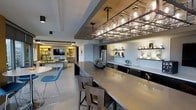 This single-floor space showcases a thoughtfully designed open-concept layout that seamlessly connects the kitchen, bar, and living areas. The kitchen and bar zone serve as the centerpiece, featuring an expansive stone-topped island with seating for multiple guests. A striking overhead wine glass rack doubles as a statement lighting fixture, suspending glassware above the island for both functional storage and dramatic visual effect. Dark lower cabinetry contrasts with light countertops and walls, while open floating shelves display curated barware and decorative accents. A built-in wine cooler and stainless steel refrigerator add to the kitchen's practical appeal. The living area, visible beyond the kitchen, features warm accent lighting within built-in shelving units that frame a wall-mounted television, creating an inviting entertainment zone. A neutral color palette of whites and grays is punctuated by pops of blue in the seating, lending a contemporary feel. Large-format floor tiles run throughout, unifying the space and enhancing its sense of openness. Recessed and track lighting provide layered illumination, complementing the overall modern aesthetic.