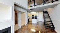 This two-story loft showcases an industrial-modern aesthetic defined by soaring double-height ceilings and richly polished concrete floors that reflect light throughout the main level. The open-concept ground floor seamlessly connects the living area to a well-appointed kitchen featuring light wood cabinetry, a central island, black appliances including a refrigerator, range, and dishwasher, and a dark granite countertop. A sliding barn door on the main level adds a stylish, functional element, providing access to an additional room or storage area. Track lighting and flush-mount ceiling fixtures ensure the space is well-lit throughout. A striking open-tread staircase with black metal railings leads to the upper mezzanine level, which is enclosed by a decorative black metal mesh railing, offering an open view down to the main floor below. This upper level provides additional versatile living or workspace. The combination of raw concrete flooring, exposed structural elements, and black metal accents throughout gives the space a cohesive, urban character that balances functionality with contemporary design.