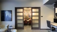 This refined single-floor space makes a strong architectural impression through its thoughtful design and upscale finishes. The centerpiece is a private meeting or dining room accessed through a pair of grand double doors, each paneled with four frosted glass insets set within dark-stained wood frames. The frosted glass provides a sense of privacy while still allowing warm, ambient light to filter into the surrounding corridor, creating an inviting glow.

Inside the enclosed room, a dramatic branching chandelier serves as the focal point, casting a warm, layered light throughout the interior. The room is furnished with a substantial central table surrounded by richly upholstered chairs in a deep reddish-brown tone, lending a sense of formality and comfort. Decorative elements, including a sculptural centerpiece and floral accents, add visual interest and sophistication.

The surrounding corridor features neutral-toned walls, carpet flooring, and clean architectural lines that complement the overall modern aesthetic. The design balances openness with privacy, making this space well-suited for professional gatherings, private dining, or executive meetings. The combination of contemporary lighting, quality materials, and refined furnishings establishes a polished and welcoming atmosphere throughout.
