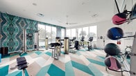 Located at 7950 Shoals Dr in Orlando, FL, this contemporary fitness center is a well-appointed, single-floor facility designed to deliver a motivating and visually dynamic workout environment. The space is immediately distinguished by its striking geometric-patterned flooring in teal, cream, and gray tones, which adds a bold design element throughout the room.

Floor-to-ceiling windows line multiple walls, flooding the interior with abundant natural light and offering sweeping views of the surrounding palm-lined landscape and outdoor pool area. A ceiling fan overhead contributes to a comfortable, airy atmosphere. A vibrant, large-scale abstract wallcovering in teal and earth tones anchors the accent wall, adding artistic depth to the space.

The fitness center is comprehensively equipped for a full-body workout. Cardio equipment includes multiple treadmills, elliptical trainers, and stationary bikes, all positioned to take full advantage of the scenic outdoor views. The strength training area features a complete dumbbell rack, medicine balls, adjustable benches, and a variety of cable and resistance machines. Wall-mounted mirrors expand the sense of space and allow for proper form monitoring. A wall-mounted flat-screen television provides entertainment during workouts. The overall ambiance is modern, energetic, and resort-inspired.