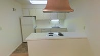 This single-floor space centers around a practical and characterful kitchen. The most striking element is the large copper range hood, which serves as a bold focal point and adds a warm metallic contrast to the otherwise neutral palette. The kitchen is designed around a peninsula-style island that houses a four-burner electric cooktop, providing a natural division between the cooking area and the rest of the space. Along the back wall, white cabinetry offers ample storage both above and below the counter, flanking a stainless steel sink with under-cabinet lighting that brightens the workspace. A full-size refrigerator is integrated into the cabinetry layout, completing the suite of appliances. The countertops are clean white surfaces that maintain a consistent, understated aesthetic throughout. Flooring transitions from a lighter surface in the kitchen area to wood-toned flooring beyond the island, subtly delineating zones within the open layout. The overall ambiance is functional and straightforward, with the copper range hood providing a distinctive design element that adds visual interest to an otherwise classic, neutral interior.