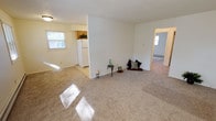 This single-floor unit offers a straightforward and functional layout, as illustrated by the overhead floor plan. The living room is notably spacious, featuring neutral-toned carpet, multiple windows that allow generous natural light to fill the room, a wall-mounted air conditioning unit, and baseboard heating — a combination that provides year-round climate control. The open flow connects the living area to the kitchen, which is separated by a partial wall. The kitchen is equipped with warm oak cabinetry extending to the ceiling for ample storage, a double sink, white appliances including a refrigerator and stove, and easy-care tile flooring.

Two carpeted bedrooms provide comfortable accommodations. One bedroom features dual windows on adjacent walls, flooding the space with natural light, while the other includes a single window and baseboard heating. Both rooms offer generous wall space suitable for a variety of furniture arrangements. An entry area features two closet or door options, providing convenient storage near the front entrance. The bathroom is finished with a full-size tub and shower combination, complemented by a light, neutral color palette throughout. Overall, the unit presents a clean, practical living environment with consistent finishes across all rooms.