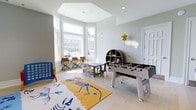 Located at 193 Tenby Chase Drive in Delran, New Jersey, this single-floor community space is thoughtfully designed to serve as a lively hub for recreation and social activity. The room is bathed in an abundance of natural light, courtesy of a striking bay window arrangement that stretches across one wall, offering views of the surrounding neighborhood and creating a warm, welcoming atmosphere throughout.

The interior features light-toned hardwood flooring and soft sage-green walls that lend the space a clean, contemporary feel. A coffered ceiling detail above the bay window area adds a touch of architectural elegance and visual depth to the room.

The space is well-appointed for entertainment and play. A full-size foosball table anchors the center of the room, making it an instant focal point for friendly competition. A vibrant, eye-catching area rug with a playful dinosaur motif adds color and personality to the floor. A colorful open bookshelf stocked with toys and games provides ample entertainment options, while a classic Connect Four game stands ready for use. A cluster of folding tables and chairs near the window bay offers a casual seating area, ideal for games, crafts, or conversation. The overall ambiance is energetic yet comfortable, making this space a versatile and enjoyable gathering place for residents of all ages.