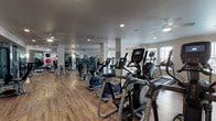 This fitness center offers a comprehensive workout environment spread across a single, open floor plan. The space features durable wood-look flooring throughout, providing a clean and modern aesthetic that complements the neutral-toned walls and recessed lighting overhead. Ceiling fans are distributed throughout the facility, ensuring consistent air circulation during workouts.

The cardio zone is well-stocked with a full lineup of Matrix-brand treadmills, elliptical trainers, stair climbers, upright bikes, and recumbent bikes, many equipped with personal display screens. Natural light filters in through several windows, creating a bright and energizing atmosphere in this section of the facility.

The strength training area is equally robust, featuring multiple cable and weight machines, adjustable benches, and a dedicated free weights section complete with a full dumbbell rack and medicine balls in various sizes. Floor-to-ceiling mirrors line the walls in the strength zone, enhancing the sense of space and allowing users to monitor their form during exercises.

Structural columns are thoughtfully integrated into the layout without disrupting the flow between zones. The overall ambiance is clean, professional, and well-maintained, making this fitness center a highly functional space suited for a wide range of fitness goals and experience levels.