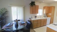 This single-story space showcases a seamlessly integrated kitchen and dining area, designed for both functionality and comfort. The kitchen features warm honey-toned wood cabinetry with generous upper and lower storage, complemented by a clean, light-colored countertop that extends to accommodate a double sink positioned beneath a window. Natural light fills the space through multiple windows dressed with vertical blinds, creating a bright and airy atmosphere throughout.

The dining area sits adjacent to the kitchen, anchored by a round glass-top table paired with dark leather chairs, offering an elegant yet practical setup for everyday meals. A large window with vertical blinds overlooks greenery outside, bringing a refreshing connection to the outdoors. The light-toned tile flooring runs throughout, contributing to the open feel of the space. Decorative touches, including potted plants atop the cabinetry and tasteful tabletop accessories, add a lived-in warmth. The neutral wall tones and consistent design elements throughout create a cohesive, welcoming environment well-suited for daily living.