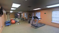 This fitness center offers a practical and functional workout environment designed to accommodate a variety of exercise routines. The space is finished with warm terracotta-toned walls and carpeted flooring, creating a comfortable atmosphere for physical activity. Recessed ceiling light panels provide consistent, even illumination throughout the room, complemented by natural light streaming in through multiple windows along the exterior wall.

The facility is outfitted with a range of cardio equipment, including a treadmill and an elliptical trainer, ideal for aerobic workouts. A multi-station strength training machine offers versatility for resistance exercises, while additional equipment such as a weight bench and exercise balls cater to a broader range of fitness needs. A large mirror spans one wall, enhancing the sense of space and allowing users to monitor their form during workouts.

A bulletin board and vending machine are visible near the entrance, adding convenience for users. Safety signage is posted on the walls, reflecting a commitment to a secure workout environment. The single-floor layout ensures easy accessibility, making this fitness center a comprehensive and user-friendly facility for everyday exercise.