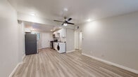This single-floor unit offers a clean, modern aesthetic defined by light-toned luxury vinyl plank flooring that runs consistently throughout the entire space, creating a cohesive and low-maintenance interior. The open-concept main area seamlessly connects the living space to the kitchen, fostering an efficient, airy layout. A ceiling fan with an integrated light fixture is centrally positioned above the living area, providing both ventilation and ambient lighting alongside recessed ceiling lights.

The kitchen is well-appointed with white shaker-style cabinetry, a classic white subway tile backsplash, dark countertops, stainless steel appliances including a gas range and microwave, and a stainless steel refrigerator. An in-unit washer is conveniently located adjacent to the kitchen. A window fitted with horizontal blinds allows natural light into the kitchen area, which also features a secondary exterior door.

The bedroom is a well-proportioned, bright room with its own ceiling fan, a window with blinds, and a dedicated exterior entry door, offering added privacy and accessibility. The bathroom presents a clean, minimalist design with white walls, a tub and shower combination, and matching light wood-toned flooring, completed with brushed nickel hardware and fixtures.