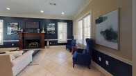 This well-appointed single-floor space offers a cohesive and sophisticated interior defined by a striking navy blue and warm beige color palette carried throughout. Crown molding and large, white-trimmed windows are consistent architectural features that lend a refined, classic character to every room.

The open-concept layout seamlessly connects the kitchen, living area, and dining spaces. The kitchen is outfitted with rich dark wood cabinetry, granite countertops, a central island with bar seating, and stainless steel appliances, including a full-size refrigerator. A glazed French door connects the kitchen to the adjacent living spaces, promoting an open and airy flow.

The living area is anchored by a traditional wood-mantel fireplace flanked by navy blue accent walls, with recessed lighting overhead and large windows dressed in white plantation shutters that allow generous natural light. Bold abstract artwork echoes the navy and gold tones found throughout the space.

The dining nook is an intimate yet stylish area featuring a round dark wood table surrounded by plush navy velvet chairs, set against a deep navy accent wall and surrounded by shuttered windows. Additional seating areas with tufted navy wingback chairs and glass side tables provide comfortable lounging spots throughout, completing a space that balances elegance with inviting warmth.