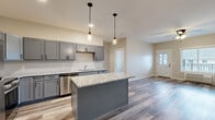 Located at 2988 Radio Rd in River Falls, WI, this 1-bedroom, 1-bathroom unit offers a thoughtfully designed layout with modern finishes throughout. The open-concept kitchen and living area serves as the heart of the space, where gray shaker-style cabinetry, granite countertops, and stainless steel appliances — including a range, microwave, refrigerator, and dishwasher — create a functional and stylish cooking environment. A central kitchen island with a matching granite surface and pendant lighting above adds both prep space and visual appeal. Hardwood-style plank flooring runs throughout the main living areas, complementing the neutral, warm-toned walls. The living room receives natural light through a window overlooking the exterior and features a ceiling fan for added comfort. The bedroom is carpeted for warmth and comfort, includes a ceiling fan, a window with exterior views, and direct access to a dedicated walk-in closet with built-in shelving. The full bathroom is finished with a gray vanity, white countertop, a large mirror, and a tub-shower combo. An additional closet near the entryway provides convenient storage. The unit presents a clean, move-in-ready aesthetic with cohesive design choices throughout.