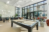 Located at 5570 Rosemead Blvd in Temple City, California, this generously sized single-floor amenity lounge is designed to foster community and leisure. The space is immediately defined by its dramatic double-height ceilings and sweeping floor-to-ceiling glass curtain walls framed in dark steel, which flood the interior with abundant natural light and provide unobstructed views of the surrounding landscaped courtyard and residential building beyond.

Light-toned hardwood-style flooring runs throughout, lending warmth and continuity to the open-plan layout. At the heart of the space sits a full-sized billiards table, complemented by a comfortable lounge area furnished with plush seating, leather ottomans, a round marble-top coffee table, and an area rug. A stylish bar-height counter and kitchen area anchors one end of the room, featuring dark cabinetry and a striking decorative wall mural as a focal backdrop. Additional casual seating clusters with bistro-style tables offer flexible spaces for socializing or working.

Large glass doors open directly to the outdoor courtyard, seamlessly blending interior and exterior living. The overall ambiance is modern, bright, and inviting, reflecting a thoughtfully curated design that balances social activity with relaxed comfort.