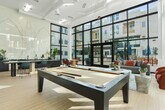 Located at 5570 Rosemead Blvd in Temple City, California, this generously sized single-floor amenity lounge is designed to foster community and leisure. The space is immediately defined by its dramatic double-height ceilings and sweeping floor-to-ceiling glass curtain walls framed in dark steel, which flood the interior with abundant natural light and provide unobstructed views of the surrounding landscaped courtyard and residential building beyond.

Light-toned hardwood-style flooring runs throughout, lending warmth and continuity to the open-plan layout. At the heart of the space sits a full-sized billiards table, complemented by a comfortable lounge area furnished with plush seating, leather ottomans, a round marble-top coffee table, and an area rug. A stylish bar-height counter and kitchen area anchors one end of the room, featuring dark cabinetry and a striking decorative wall mural as a focal backdrop. Additional casual seating clusters with bistro-style tables offer flexible spaces for socializing or working.

Large glass doors open directly to the outdoor courtyard, seamlessly blending interior and exterior living. The overall ambiance is modern, bright, and inviting, reflecting a thoughtfully curated design that balances social activity with relaxed comfort.