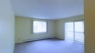 This single-floor residence offers a straightforward and functional layout designed for comfortable everyday living. The living area is the focal point of the space, characterized by soft wall-to-wall carpeting and light-toned walls that contribute to a clean, neutral aesthetic. Natural light fills the room through a large window dressed with vertical blinds, allowing for adjustable brightness throughout the day. A sliding glass door provides direct access to the outdoors, enhancing the sense of openness and connectivity to the exterior. A baseboard heating unit runs along the exterior wall, ensuring efficient and consistent warmth during cooler months. The textured ceiling adds a subtle architectural detail to the overall interior. The room's open, unobstructed layout offers flexibility for a variety of furniture arrangements, making it adaptable to different living needs. The combination of ample natural light, neutral finishes, and practical features creates a calm and livable environment suited for a range of occupants.