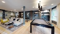 This thoughtfully designed community room offers a dynamic, open-plan layout that seamlessly blends recreation, relaxation, and functionality. Light hardwood flooring runs throughout, unifying the various zones and complementing the deep slate-blue accent walls adorned with contemporary artwork.

The game area is a standout feature, equipped with a full-size pool table and a shuffleboard table, both finished in sleek dark frames. Dramatic pendant lighting overhead adds an upscale touch to the space. Nearby, a generous lounge area is anchored by a floor-to-ceiling wood-clad feature wall housing a wide linear fireplace and a large mounted flat-screen TV, creating an inviting focal point. Comfortable sectional sofas, accent chairs in bold yellow, and layered area rugs complete the cozy yet modern atmosphere.

The catering kitchen is fully appointed with stainless steel appliances, a cooktop with a professional range hood, wall ovens, white cabinetry, and a long communal dining table beneath sculptural bronze pendant lights — ideal for gatherings and events.

Glass-enclosed private rooms along one side of the space provide flexible areas for meetings or focused use. Large windows invite natural light and offer views of the surrounding greenery, further enhancing the welcoming ambiance of this versatile community space.