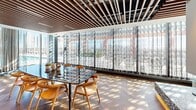 Located in the heart of Miami's vibrant Wynwood neighborhood at 55 NW 29th St, this thoughtfully designed single-floor space combines refined materials with an open, modern layout. Rich herringbone-patterned hardwood flooring flows throughout, anchoring the space with warmth and visual interest. Above, a striking wood slat ceiling adds architectural depth and a natural, textural contrast against the exposed black ceiling infrastructure beyond — a design choice that merges industrial and organic elements seamlessly.

Floor-to-ceiling sheer curtain panels line an entire wall of expansive windows, bathing the interior in abundant natural light and offering views of an outdoor terrace and pool beyond. The interplay of sunlight through the curtains creates a dynamic, ever-shifting atmosphere throughout the day.

A sleek kitchenette anchors one end of the space, featuring warm wood cabinetry, a bold Calacatta marble backsplash, and an integrated sink — a functional yet visually striking focal point. A statement dining table with a dramatic dark marble top and natural wood chairs provides an elegant gathering area. A white partition wall creates subtle spatial definition without closing off the open-plan flow. Together, these elements create a polished, inviting environment well-suited for professional or social use.