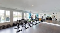 This fitness center is designed to support a wide range of workout routines within a bright, contemporary setting. The space is organized into distinct functional zones, beginning with a dedicated cycling studio lined with professional-grade spin bikes arranged in rows. Large windows along one wall flood the area with natural light while offering views of an outdoor pool and surrounding grounds, creating an energizing atmosphere during workouts.

Adjacent to the cycling area, a well-stocked strength and functional training zone houses a comprehensive selection of kettlebells, dumbbells arranged on tiered racks, stability balls, resistance bands, and exercise mats. Floor-to-ceiling mirrors line one wall, visually expanding the space and allowing for form monitoring during exercise.

A central check-in or reception counter with open shelving and pendant lighting serves as a hub between zones, offering a polished, organized point of entry. Beyond this, a carpeted cardio area features treadmills and other upright machines, distinguished by its vaulted ceiling with exposed dark beams that add architectural character.

A dedicated open studio area with dark hardwood-style flooring, a wall-mounted flat-screen television with a soundbar, and framed artwork provides a versatile space for stretching, yoga, or group classes. Recessed lighting throughout ensures consistent illumination across all zones.