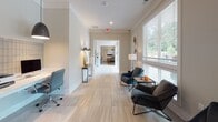 This well-appointed single-floor office space presents a clean, contemporary aesthetic defined by a neutral palette of whites, warm grays, and soft greiges. Light-toned large-format tile flooring runs throughout, contributing to an open and airy atmosphere.

The business center area features a sleek, built-in marble-topped workstation along a tiled accent wall, equipped with desktop computers and a printer, illuminated by a pair of pendant lights with textured gray shades. The setup is functional and refined, designed to accommodate multiple users simultaneously.

Adjacent to the workstation, a welcoming lounge area offers modern upholstered chairs in deep charcoal blue arranged around a compact side table. This seating nook is positioned alongside a bank of large windows dressed with white horizontal blinds, allowing generous natural light to filter in while providing views of the surrounding greenery.

A corridor connects the various areas of the space, lined with subtle wall sconces and a console table that add warmth and dimension. Curated framed artwork, including a bold still-life canvas, adds artistic character to the walls. The overall ambiance is professional yet inviting, balancing functionality with tasteful design.