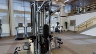 This well-appointed two-story fitness center offers a comprehensive and thoughtfully designed workout environment. The main floor is expansive and open, featuring a wide selection of strength training equipment, including a multi-station cable machine, a full dumbbell rack with adjustable benches, and additional resistance machines positioned throughout the space. The cardio zone is equally equipped, with treadmills, elliptical trainers, and other aerobic machines arranged near large windows that invite natural light and provide an airy atmosphere.

The facility's most striking architectural feature is its soaring double-height ceiling, which creates a dramatic sense of openness and volume. A mezzanine level overlooks the main floor, accessible via a staircase and bordered by sleek glass railings that maintain visual connectivity between the two levels. Warm wood-toned accents along the upper balcony contrast tastefully with the clean white walls and modern linear lighting throughout.

Decorative touches such as framed artwork and lush indoor plants add warmth and visual interest to the space. The flooring is designed for durability and safety, appropriate for high-traffic fitness use. Abundant overhead lighting, combined with natural light from large windows, ensures the entire facility is bright and welcoming at all hours.
