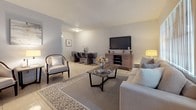 This single-story residence is designed with a clean, contemporary aesthetic, utilizing a neutral palette of whites and soft grays to create a bright and cohesive atmosphere throughout. Light tile flooring runs consistently across all areas, reinforcing the open and airy feel of the space.

The living area is generously sized, accommodating a sectional sofa, accent chair, and a rustic wood coffee table atop a patterned area rug. Vertical blinds on a large window allow for adjustable natural light, while a media console with a wall-mounted television anchors one side of the room. The living space flows seamlessly into a dedicated dining area, which connects directly to the kitchen.

The kitchen features rich dark wood cabinetry paired with granite-style countertops, a stainless steel range with overhead ventilation, and a tile backsplash, offering a functional and stylish cooking environment.

The primary bedroom is a standout feature, offering generous proportions, serene light tones, and a full glass sliding door that opens to a view of lush green outdoor surroundings. The room includes closet space and connects to a dedicated workspace area. The bathroom presents a dark wood vanity with a white basin, a framed mirror, and tile flooring, maintaining the consistent style found throughout the residence.