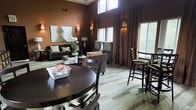 This single-floor space offers a sophisticated, welcoming atmosphere defined by its notably high ceilings and rich, earth-toned interior design. The walls are finished in deep warm browns, complemented by strategically placed wall sconces that cast a soft, ambient glow throughout the room. Large windows dressed with floor-length draping curtains allow natural light to filter in, offering views of surrounding greenery and enhancing the space's airy feel despite its cozy aesthetic.

The layout is thoughtfully divided into distinct functional zones. A casual lounge area features a dark upholstered sofa and coordinating accent chairs arranged around a glass-topped coffee table, creating an inviting conversational setting. Nearby, a round dark wood dining table with matching chairs provides a more structured seating option, suitable for casual dining or social gatherings. A counter-height bar table with tall chairs occupies a separate corner, adding a relaxed, social dimension to the space.

The flooring consists of a patterned carpet in neutral tones that ties the various zones together cohesively. Decorative artwork, fresh floral arrangements, and layered lighting contribute to the polished yet comfortable ambiance throughout.