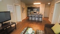 This single-floor residence showcases an open-concept design that allows the living, dining, and kitchen areas to flow naturally into one another. Rich hardwood flooring runs throughout the main living space, adding warmth and visual continuity across the layout.

The kitchen is equipped with stainless steel appliances, including a refrigerator, microwave, and range, complemented by dark espresso cabinetry that provides a sleek, modern contrast to the warm-toned walls and flooring. A peninsula-style counter serves as a casual dining area with seating for several bar-height chairs, making it a functional and social hub of the space.

The living area offers comfortable space for seating arrangements, with a coffee table and area rug grounding the room. Neutral beige walls provide a versatile backdrop throughout. Recessed ceiling vents and overhead lighting ensure the space is well-ventilated and evenly lit.

Multiple interior doors are visible, suggesting additional private rooms such as a bedroom and bathroom. White panel doors and clean baseboards contribute to the overall tidy, well-maintained aesthetic of the residence.