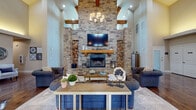 This single-floor space makes an immediate impression with its dramatic architectural details and thoughtful design. The centerpiece of the main living area is a floor-to-ceiling stacked-stone fireplace, flanked by built-in firewood storage niches and crowned with a substantial wooden mantel. Exposed wooden ceiling beams add warmth and rustic character, while the soaring ceiling height creates an expansive, airy atmosphere. A statement chandelier hangs above the seating area, complementing the rich hardwood flooring that flows throughout the space. The living area is generously proportioned, comfortably accommodating multiple seating arrangements around a large area rug.

The open-concept kitchen features granite countertops, rich dark wood cabinetry, stainless steel appliances, and a wall-mounted range hood. An extended breakfast bar with corbel supports provides casual seating for several barstools, illuminated by a trio of industrial-style pendant lights. A pass-through design connects the kitchen seamlessly to the adjoining living and dining spaces.

Additional areas of the space include a welcoming seating nook with cove lighting, a casual dining area, and a lounge zone with direct access to an outdoor area through glass-panel doors. Large windows throughout invite abundant natural light, enhancing the warm, inviting ambiance of the entire space.