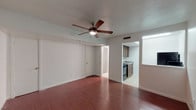 This single-floor unit offers a straightforward, functional layout centered around an open main living area. The space is finished with rich, dark hardwood-style flooring that extends throughout the main area, creating a warm and cohesive look. A five-blade ceiling fan with an integrated light fixture is centrally mounted, providing both airflow and ambient lighting to the room.

The front entry features a paneled white door with a narrow sidelight window, allowing natural light to filter into the living space. White walls throughout keep the interior feeling bright and clean, while white baseboards provide a crisp contrast to the darker flooring.

The kitchen is situated adjacent to the main living area and transitions to light ceramic tile flooring. It is equipped with dark walnut-toned cabinetry offering ample upper and lower storage, a granite-style countertop with a stainless steel sink, and a black gas range. A pass-through or window opening connects the kitchen visually to a secondary area, enhancing the sense of openness. Ceiling vents indicate a central HVAC system is in place. The layout is compact yet practical, making efficient use of the available space.