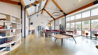 Located at 1562 Magnolia Dr in Oxford, Ohio, this expansive single-story community clubhouse offers a generously sized open-plan interior designed for socializing and recreation. The architectural centerpiece is the dramatic vaulted ceiling supported by striking exposed wood and steel beam trusses, which lend the space an industrial-rustic character. Polished concrete floors run throughout, complementing the white shiplap walls and large floor-to-ceiling windows that flood the interior with abundant natural light and offer views of the surrounding outdoors.

The main gathering area is anchored by a floor-to-ceiling stacked stone fireplace flanked by comfortable seating, including leather sofas, accent chairs, and vibrant area rugs that define a cozy lounge zone. A bar counter with stylish seating provides an additional social hub. The games area is well-appointed with a billiards table, ping-pong table, and shuffleboard, while open shelving displays curated décor. A dining and café-style seating section features bistro tables, wall-mounted televisions, and warm sconce lighting. Pendant chandeliers and industrial-style hanging fixtures enhance the ambiance throughout. Exposed ductwork adds to the space's modern industrial aesthetic, creating a dynamic and inviting environment for community gatherings.