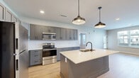 This well-appointed 2-bedroom, 2-bathroom single-story residence offers a clean, modern aesthetic defined by light-toned hardwood flooring, neutral wall colors, and consistent matte black hardware accents throughout. The open-concept kitchen is a standout feature, showcasing gray shaker-style cabinetry with ample upper and lower storage, white subway tile backsplash, stainless steel appliances including a refrigerator, electric range, and built-in microwave, and a large center island with a white quartz countertop and undermount sink, illuminated by two pendant lights. The kitchen flows seamlessly into the dining and living areas, where large windows and a glass door invite abundant natural light and outdoor views. The primary bedroom is generously sized and accompanied by a spacious walk-in closet with wire shelving systems. The second bedroom offers a well-proportioned layout with the same consistent flooring. Both bathrooms are finished with gray vanities, white countertops, and a tub/shower combination clad in white subway tile with matte black fixtures. Additional functional spaces include a dedicated laundry room, utility room, hallway, and three closets, ensuring excellent storage and practicality throughout.