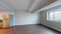 This single-floor residence offers a thoughtfully designed layout that balances open living spaces with comfortable private areas. The open-concept living and dining area is finished with rich wood-tone flooring and neutral grey walls, creating a bright and welcoming atmosphere. Architectural beam details along the ceiling add visual interest and a subtle structural character to the space.

The kitchen is a standout feature, offering a complete suite of stainless steel appliances including a refrigerator, electric range, over-the-range microwave, and dishwasher. White arched cabinetry provides generous storage, while dark granite countertops and a classic white subway tile backsplash with under-cabinet lighting round out the space with a clean, polished aesthetic. A partial wall separates the kitchen from the living area, maintaining an open feel while defining each zone.

The bedroom is generously sized, carpeted for comfort, and features a large window that fills the room with natural light. Dual arched bi-fold closet doors offer ample storage. The full bathroom includes a soaking tub and shower combination with white tile surround, white tile flooring, a granite-topped vanity, and clean, neutral finishes throughout. A hallway with additional storage connects the rooms, completing this well-organized floor plan.