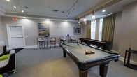 This well-appointed game room offers a refined yet relaxed atmosphere designed for social gatherings and recreational play. The centerpiece of the space is a full-sized billiards table with a sage green felt surface, complemented by a shuffleboard table positioned along the far wall. Pendant lighting hangs gracefully above the billiards table, casting a warm, focused glow, while recessed lighting and a track lighting system illuminate the rest of the room evenly. Crown molding runs along the perimeter of the ceiling, adding a touch of classic elegance to the space. Neutral-toned carpeting covers the floor, contributing to a comfortable and inviting feel underfoot. Along one wall, tall bistro-style tables paired with high-backed chairs provide a casual seating area, ideal for spectators or conversation. Natural light filters into the room through windows dressed with warm gold drapery, brightening the space during the day. Contemporary artwork adorns the walls, enhancing the polished aesthetic. The overall ambiance strikes a balance between a sophisticated lounge and a fun, activity-focused environment, making it a versatile and welcoming destination for leisure and socializing.