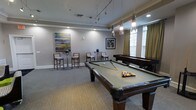 This well-appointed game room offers a refined yet relaxed atmosphere designed for social gatherings and recreational play. The centerpiece of the space is a full-sized billiards table with a sage green felt surface, complemented by a shuffleboard table positioned along the far wall. Pendant lighting hangs gracefully above the billiards table, casting a warm, focused glow, while recessed lighting and a track lighting system illuminate the rest of the room evenly. Crown molding runs along the perimeter of the ceiling, adding a touch of classic elegance to the space. Neutral-toned carpeting covers the floor, contributing to a comfortable and inviting feel underfoot. Along one wall, tall bistro-style tables paired with high-backed chairs provide a casual seating area, ideal for spectators or conversation. Natural light filters into the room through windows dressed with warm gold drapery, brightening the space during the day. Contemporary artwork adorns the walls, enhancing the polished aesthetic. The overall ambiance strikes a balance between a sophisticated lounge and a fun, activity-focused environment, making it a versatile and welcoming destination for leisure and socializing.