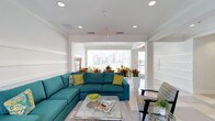 This single-floor space is designed as a stylish and inviting communal lounge, offering a well-appointed environment ideal for relaxation and socializing. The interior is anchored by a bold teal sectional sofa accented with yellow and patterned throw pillows, creating a lively and contemporary focal point. A sleek glass-and-chrome coffee table sits at the center, complemented by additional seating that adds to the room's flexible layout.

The décor throughout is fresh and vibrant, with potted tropical plants and colorful floral arrangements introducing natural elements that enhance the cheerful atmosphere. The walls feature classic wainscoting details and are finished in a clean, crisp white, contributing to the space's bright and airy feel.

Recessed lighting is evenly distributed across the ceiling, ensuring consistent illumination throughout the day, while large windows along the far wall allow generous amounts of natural light to pour in. These windows also frame views of a landscaped outdoor area, connecting the interior to its surrounding environment.

The flooring is finished in light-toned tile, reinforcing the open and clean aesthetic of the space. An open passageway leads seamlessly into an adjacent area, further enhancing the sense of spaciousness and flow throughout the layout. Overall, the space strikes a balance between comfort and modern design.