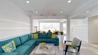 This single-floor space is designed as a stylish and inviting communal lounge, offering a well-appointed environment ideal for relaxation and socializing. The interior is anchored by a bold teal sectional sofa accented with yellow and patterned throw pillows, creating a lively and contemporary focal point. A sleek glass-and-chrome coffee table sits at the center, complemented by additional seating that adds to the room's flexible layout.

The décor throughout is fresh and vibrant, with potted tropical plants and colorful floral arrangements introducing natural elements that enhance the cheerful atmosphere. The walls feature classic wainscoting details and are finished in a clean, crisp white, contributing to the space's bright and airy feel.

Recessed lighting is evenly distributed across the ceiling, ensuring consistent illumination throughout the day, while large windows along the far wall allow generous amounts of natural light to pour in. These windows also frame views of a landscaped outdoor area, connecting the interior to its surrounding environment.

The flooring is finished in light-toned tile, reinforcing the open and clean aesthetic of the space. An open passageway leads seamlessly into an adjacent area, further enhancing the sense of spaciousness and flow throughout the layout. Overall, the space strikes a balance between comfort and modern design.