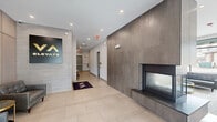 The VA Elevate Lobby and Fireside Lounge is a thoughtfully designed communal space spread across a single floor, offering a welcoming and sophisticated environment for residents and guests. The entry corridor features large-format light stone tile flooring, a bold branded wall sign, and a dramatic floor-to-ceiling dark tile fireplace surround that anchors the space with a striking architectural focal point. A granite hearth and sleek modern firebox add warmth and visual interest to the entry experience. Connected hallways provide access to elevators and stairwells, maintaining efficient circulation throughout. The fireside lounge area transitions into a bright, open living room furnished with tufted leather sofas, upholstered armchairs, and a gold-accented glass coffee table atop a layered area rug. A wall-mounted large-screen television is integrated into a built-in cabinetry unit featuring white lower cabinets, warm wood upper storage, and a wet bar with a sink and beverage refrigerator. Floor-to-ceiling black-framed windows flood the lounge with abundant natural light and provide unobstructed views of the surrounding streetscape. The overall aesthetic is contemporary and refined, combining neutral tones, natural wood accents, and polished finishes.