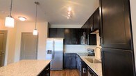 Located at 211 River Landing Dr. on Daniel Island, SC, this single-floor residence offers a thoughtfully designed living space with modern finishes throughout. The kitchen is a standout feature, showcasing rich dark espresso cabinetry that extends to the ceiling, providing ample storage and a sleek, contemporary aesthetic. Light granite countertops offer a striking contrast against the dark cabinetry, while a tile backsplash adds a refined finishing touch. The kitchen is equipped with a full suite of stainless steel appliances, including a French door refrigerator with an in-door water and ice dispenser, a built-in microwave, and a range. A deep undermount sink sits beneath a modern faucet, and pendant lighting adds warmth and visual interest above the countertop island or bar area. Track lighting illuminates the space efficiently, highlighting the kitchen's clean lines and high-quality materials. The warm hardwood-style flooring runs throughout, tying the space together with a cohesive and inviting feel. The overall ambiance is polished and contemporary, combining functional design with upscale finishes that make this Daniel Island residence both practical and visually appealing.