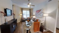 This single-story living space is thoughtfully designed to balance comfort and style. The room features rich hardwood flooring that extends throughout, providing a warm and grounding foundation. A ceiling fan with an integrated light fixture is centrally mounted, offering both functionality and a subtle decorative element.

Natural light enters through a large window dressed with floor-length patterned drapes, creating a bright and welcoming atmosphere. The seating area is anchored by a plush area rug and centers around a glass-top coffee table with a mirrored frame, complemented by a coordinating side table. A neutral-toned sofa is styled with gray and burnt orange throw pillows, echoed by a matching armchair arrangement near the window.

A bold figurative canvas painting adds an artistic focal point to the wall, while a dark-finished media console along the opposite wall provides storage and contrast. Layered lighting from multiple floor and table lamps contributes to the room's warm, inviting ambiance, making this a well-appointed and visually cohesive living area.