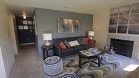 This single-floor space makes a strong design statement from the moment you step inside. The living area serves as the centerpiece, anchored by a deep charcoal accent wall that provides a bold, sophisticated backdrop. A large, colorful abstract canvas painting adds a dynamic focal point, drawing the eye and infusing the room with warmth and artistic energy. The space is furnished with a navy blue sofa adorned with a mix of patterned and solid throw pillows in burnt orange, black-and-white, and cream tones, complemented by an orange throw blanket for a pop of color. An oval glass-top coffee table sits atop a striking wavy, multicolored area rug in shades of blue, green, and gold, adding visual interest and texture to the floor. A round striped pouf provides additional casual seating. Matching side tables with elegant table lamps flank the sofa, casting warm, ambient light throughout the room. The neutral carpeted flooring and lighter surrounding walls balance the drama of the accent wall, creating a cohesive and inviting atmosphere. The overall ambiance is modern and thoughtfully curated, blending bold color choices with comfortable furnishings to create a space that is both visually engaging and welcoming.