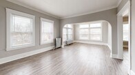 Located at 12933 Clifton Blvd in Lakewood, Ohio, this 2-bedroom, 1-bathroom residence showcases a blend of classic character and functional design across a well-organized single floor. The spacious living room immediately impresses with its crown molding, neutral gray walls, light wood-style flooring, and an elegant arched passageway flanked by large multi-pane windows that flood the room with natural light. Vintage cast-iron radiators are present throughout, adding a charming period detail while providing reliable warmth. The separate dining room offers additional living space with its own natural light and flows conveniently into the kitchen. The kitchen is equipped with warm oak cabinetry, dark countertops, a subway tile backsplash, a double sink, and a full-size refrigerator, with a door providing direct exterior access. Both bedrooms are comfortably sized, featuring crown molding, neutral tones, and their own windows with neighborhood views — one with hardwood-style flooring and one with plush carpet. Two closets offer ample storage. The bathroom includes a full soaking tub with shower and a window for natural ventilation. A connecting hallway ties all rooms together for a practical, cohesive layout.