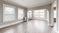 Located at 12933 Clifton Blvd in Lakewood, Ohio, this 2-bedroom, 1-bathroom residence showcases a blend of classic character and functional design across a well-organized single floor. The spacious living room immediately impresses with its crown molding, neutral gray walls, light wood-style flooring, and an elegant arched passageway flanked by large multi-pane windows that flood the room with natural light. Vintage cast-iron radiators are present throughout, adding a charming period detail while providing reliable warmth. The separate dining room offers additional living space with its own natural light and flows conveniently into the kitchen. The kitchen is equipped with warm oak cabinetry, dark countertops, a subway tile backsplash, a double sink, and a full-size refrigerator, with a door providing direct exterior access. Both bedrooms are comfortably sized, featuring crown molding, neutral tones, and their own windows with neighborhood views — one with hardwood-style flooring and one with plush carpet. Two closets offer ample storage. The bathroom includes a full soaking tub with shower and a window for natural ventilation. A connecting hallway ties all rooms together for a practical, cohesive layout.