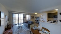 This well-appointed single-floor residence offers an open-concept layout that seamlessly connects the living, dining, and kitchen areas into one cohesive and functional space. The living area is anchored by a stylish sofa set atop a patterned area rug, complemented by a glass-top coffee table and a sleek arc floor lamp. Large sliding glass doors flood the room with natural light and lead out to a private balcony, offering sweeping panoramic views of the surrounding landscape. A modular open shelving unit serves as both a display space and a subtle divider between the living and work zones, adding visual interest and storage. The dining area features a warm-toned round table set with elegant place settings, creating an inviting space for meals and entertaining. The kitchen, partially visible from the main living area, is neatly integrated into the floor plan. Throughout, light hardwood-style flooring and neutral wall tones create a calm, contemporary ambiance. The overall design balances comfort and sophistication, making it an ideal space for both relaxing and hosting.