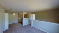 This single-floor space features an open-concept layout that connects the living area to the kitchen, creating a cohesive and functional floor plan. The living area is finished with wall-to-wall carpet in a neutral tone, contributing to a calm and understated ambiance. A ceiling fan is installed overhead, providing both practical airflow and a modest lighting source for the space.

A half-wall partition separates the living area from the kitchen, maintaining visual openness while defining each zone. The kitchen is equipped with light wood cabinetry that extends to the upper walls, offering generous storage. A pass-through window or opening in the partition enhances the connection between the two areas, making the layout feel airy and communicative.

The walls are painted in a soft, neutral gray-beige tone throughout, lending consistency and a clean look to the interior. A doorway visible on the far side of the space leads to an additional room, suggesting further utility beyond the main living and kitchen zones. The textured ceiling and understated trim work complete the straightforward, practical character of this space.