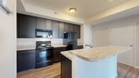 Located at 7220 E. Broad in Columbus, Ohio, this single-floor, 2-bedroom, 2-bathroom unit offers a well-organized layout designed for comfortable everyday living. The open-concept kitchen and living area serves as the heart of the home, where a peninsula with light granite-style countertops and a built-in sink doubles as a breakfast bar. Dark espresso cabinetry reaches toward the ceiling, providing generous storage, while black appliances — including a range, microwave, and refrigerator — complete the kitchen. Wood-look flooring runs throughout the main living and kitchen areas, adding warmth and durability. The living space is bright, with large windows and a sliding glass door that opens to a private balcony. Both bedrooms are carpeted and well-proportioned, with the primary bedroom offering notable square footage and outdoor-facing windows. Six closets distributed throughout the unit ensure abundant storage. The two bathrooms feature tile flooring, dark vanities with matching granite-style countertops, and clean, neutral finishes. Additional conveniences include a dedicated laundry area and a utility room. Neutral wall tones and consistent finishes throughout create a cohesive and move-in-ready atmosphere.