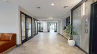 Located at 215 Fairmount Avenue in Lynn, Massachusetts, this well-appointed amenity space offers residents a refined and functional environment across a single floor. The layout encompasses four distinct areas, each designed with modern aesthetics and practical utility in mind.

The lobby serves as a polished point of entry, featuring large-format stone-look tile flooring, recessed lighting, and black-framed glass doors that lend a contemporary edge. A sleek elevator with warm wood slat detailing and a built-in credenza with a coffee station add convenience and character to the arrival experience.

Adjacent to the lobby, a generously sized lounge area provides an inviting space for relaxation and casual gatherings. A large wraparound cognac leather sectional anchors the room against a textured blue-grey accent wall with stylish wall sconces. An open shelving unit, natural light from nearby windows, and a small café-style dining area with round table seating complete this comfortable setting.

The fitness center is well-equipped with a full range of cardio machines and strength training equipment, including a Precor cable machine, free weights, and exercise accessories. Light hardwood-style flooring, blue accent walls, floor-to-ceiling mirrors, and natural light from large windows create a bright and energizing workout environment. Together, these spaces deliver a cohesive, upscale amenity experience.