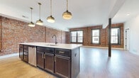 Located in the heart of downtown Columbus, this thoughtfully designed 3-bedroom, 3-bathroom residence masterfully blends historic industrial character with contemporary finishes. Exposed red brick walls are a defining architectural feature throughout, lending warmth and authenticity that contrast beautifully with crisp white walls and light hardwood flooring found across the main living areas.

The open-concept kitchen and living room serve as the centerpiece of the home. The kitchen is outfitted with dark espresso cabinetry accented by brushed gold hardware, a large center island with a white quartz countertop, a subway tile backsplash, stainless steel appliances, and a matte black sink fixture. Overhead, a row of antique brass globe pendant lights adds an industrial-chic ambiance. Large dark-framed windows allow natural light to pour in while offering views of the surrounding cityscape.

The three bedrooms offer a quieter retreat, several featuring the same striking exposed brick walls paired with soft carpet flooring. The generously sized primary bedroom provides ample space for comfortable living. Each of the three bathrooms is finished with dark wood vanities, white quartz countertops, matte black fixtures with gold accents, and glass-enclosed showers, maintaining a cohesive modern aesthetic throughout. A connecting hallway ties all rooms together seamlessly.