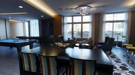 This sophisticated single-floor space seamlessly blends a dining area with a relaxed lounge, creating a versatile and inviting environment. The dining section features a sleek, dark-finished table paired with striking teal and black striped high-back chairs, adding a bold design element to the space. A decorative bowl centerpiece with orb accents anchors the table with an elegant touch.

Overhead, a contemporary ceiling fan with a metallic finish serves as a functional focal point, complemented by warm cove lighting that runs along the ceiling perimeter, casting a soft amber glow throughout. The combination of ambient and natural light creates a layered, welcoming atmosphere.

Large, floor-to-ceiling windows draped with sheer white curtains dominate one wall, flooding the interior with natural light and offering views of the surrounding urban streetscape. The lounge area beyond features plush neutral-toned seating arranged around a low coffee table, ideal for casual gatherings.

Dark wood accent walls and rich, moody tones contrast with the lighter architectural elements, giving the space a refined, contemporary character. A large-scale artwork adds a dramatic visual element in the background. The overall ambiance is modern and polished, balancing comfort with sophisticated design.