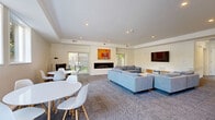 Located at 703 Whitney Avenue in New Haven, CT, this single-floor space offers a thoughtfully designed open-plan living room that makes excellent use of its generous footprint. The interior features a clean, modern aesthetic defined by crisp white walls, recessed lighting, and a tray ceiling that adds architectural interest and a sense of depth to the room.

The living area is anchored by a sleek linear fireplace flanked by large sliding glass doors that flood the space with natural light and provide direct views of the private outdoor area. A wall-mounted flat-screen TV and a contemporary media console complement the seating arrangement, which includes a plush sectional sofa and accent chairs arranged around a marble-top coffee table. Patterned wall-to-wall carpeting in a neutral blue-grey tone adds warmth and texture throughout.

A dedicated dining area with a round white table and modern chairs offers a distinct yet open zone for meals and gatherings. Colorful artwork adorns the walls, contributing vibrancy to the otherwise understated palette.

The space extends outdoors to a fenced backyard shaded by mature trees, complete with a gas grill and patio area — ideal for outdoor entertaining. The combination of modern finishes, natural light, indoor-outdoor connectivity, and thoughtful layout makes this a highly functional and inviting space.