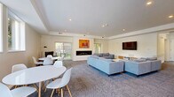 Located at 703 Whitney Avenue in New Haven, CT, this single-floor space offers a thoughtfully designed open-plan living room that makes excellent use of its generous footprint. The interior features a clean, modern aesthetic defined by crisp white walls, recessed lighting, and a tray ceiling that adds architectural interest and a sense of depth to the room.

The living area is anchored by a sleek linear fireplace flanked by large sliding glass doors that flood the space with natural light and provide direct views of the private outdoor area. A wall-mounted flat-screen TV and a contemporary media console complement the seating arrangement, which includes a plush sectional sofa and accent chairs arranged around a marble-top coffee table. Patterned wall-to-wall carpeting in a neutral blue-grey tone adds warmth and texture throughout.

A dedicated dining area with a round white table and modern chairs offers a distinct yet open zone for meals and gatherings. Colorful artwork adorns the walls, contributing vibrancy to the otherwise understated palette.

The space extends outdoors to a fenced backyard shaded by mature trees, complete with a gas grill and patio area — ideal for outdoor entertaining. The combination of modern finishes, natural light, indoor-outdoor connectivity, and thoughtful layout makes this a highly functional and inviting space.