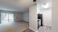This single-floor unit features a practical, open-concept layout where the living area flows directly into the kitchen. The living space is finished with neutral-toned carpet and painted walls, and is equipped with a wall-mounted air conditioning unit. A sliding glass door provides access to the exterior, allowing natural light into the room. A ceiling fan overhead adds functionality to the space.

The kitchen is efficiently designed in a galley configuration, featuring white shaker-style cabinetry with black hardware, light granite-patterned countertops, wood-look vinyl plank flooring, and black appliances including a refrigerator, range, and dishwasher. Ample upper and lower cabinet storage is present throughout.

The bedroom is a generously sized, carpeted room with neutral gray walls, baseboard heating units, and a window offering views of surrounding greenery. A six-panel door leads to an adjoining area with additional closet access.

The bathroom includes a full-size soaking tub with an integrated white surround, overhead shower, chrome fixtures, and wood-look flooring. The unit maintains a clean, neutral palette throughout, offering a functional and move-in ready living environment.